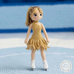 May include: Crocheted doll of a figure skater wearing a gold dress and white ice skates. The doll has blonde hair in a ponytail and is standing on a blue and white surface. The doll is handmade.