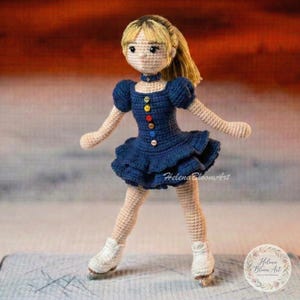 May include: A handmade crochet doll in a skating outfit. The doll has blonde hair and is wearing a navy blue dress with colorful buttons. The doll is wearing white ice skates and is posed as if skating on ice.