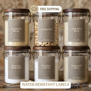 May include: Six clear glass storage canisters with dark wood lids and metal latch closures. Each jar features a rectangular taupe label with white text reading Almond Meal, Almonds, Arborio Rice, Baking Powder, Baking Soda, and Basmati Rice.