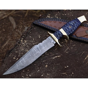 Puede incluir: Un cuchillo de caza de acero de Damasco con una hoja texturizada y un mango azul y dorado. El cuchillo tiene una funda de cuero con un patrón decorativo. El cuchillo está sobre una superficie de madera.