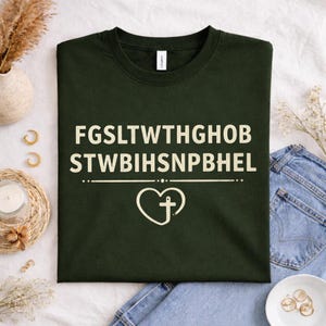 May include: Dark green t-shirt with white text and a heart-shaped design with a cross. The text reads "FGSLT WTHGHOB STWBIHSNPBHEL". The shirt is folded on a white surface with accessories in the background.