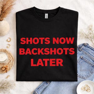 May include: Black t-shirt with the red text "SHOTS NOW BACKSHOTS LATER". The shirt is displayed on a white surface with a candle, jewelry, and denim jeans.