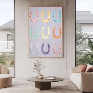 May include: A framed print displaying nine horseshoes in a grid, each in a different pastel shade. The colours include pink, yellow, blue, green, orange, purple, and white, set against a pale grey backdrop. The artwork is displayed in a contemporary living room.