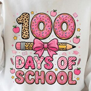 Puede incluir: Sudadera blanca con un gráfico colorido que celebra los 100 días de escuela. El diseño incluye el número "100" con un "1" con estampado de leopardo, rosquillas rosas, un lápiz, un lazo rosa, manzanas, estrellas y las palabras "DAYS OF SCHOOL".