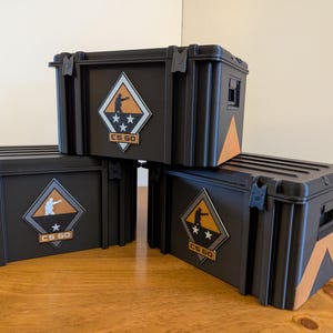 May include: Three black storage boxes with orange and white accents. Each box features a diamond-shaped emblem with a silhouette of a person aiming a gun, stars, and the text "CS GO". The boxes are stacked on a wooden surface.