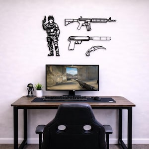 May include: Black metal wall art featuring a soldier figure, a rifle, a pistol, and a curved knife. A computer monitor displays a video game scene. The desk has a keyboard, mouse, and a small figurine.
