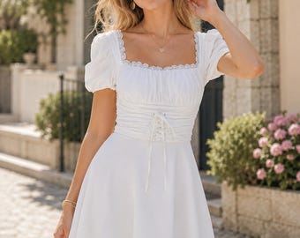May include: A white mini dress featuring a square neckline with delicate lace trim, short puff sleeves, and a structured bodice with a central corset-style lace-up detail. The skirt is flared and falls above the knee, crafted from a solid white fabric.