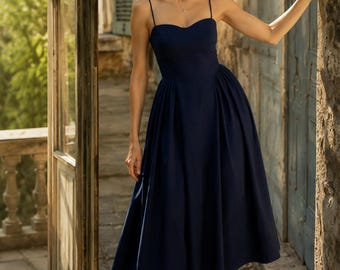 May include: A navy blue midi dress with spaghetti straps and a sweetheart neckline. The dress has a fitted bodice and a flared skirt. The person is wearing beige heels. The background includes a stone wall and a wooden door.