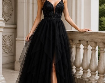 May include: A black evening gown with a plunging neckline and floral embellishments. The dress features a layered tulle skirt with a high slit, and spaghetti straps. The woman is standing on a balcony with a stone balustrade.