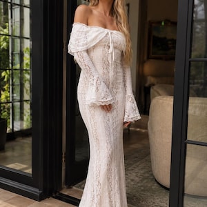 May include: A floor-length off-the-shoulder dress made of white lace. The garment features long sleeves with flared cuffs, a fitted bodice, and a tie detail at the center of the neckline. The lace fabric has a detailed floral pattern throughout.