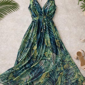 Puede incluir: Un vestido largo sin mangas con un escote en V profundo y un cierre de lazo estilo halter en el cuello. El vestido est&aacute; hecho de una tela ligera con un estampado integral de hojas de palmera en verde oscuro, verde claro y amarillo.