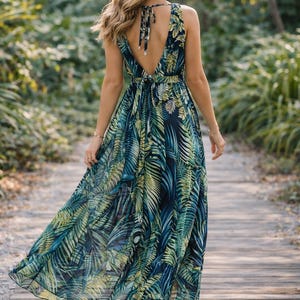 Puede incluir: Vestido largo sin mangas con estampado de hojas tropicales en tonos azules y verdes. El vestido tiene un escote en V pronunciado y un lazo en la espalda. La persona lleva tacones beige y camina por una pasarela de madera. El vestido es fluido.