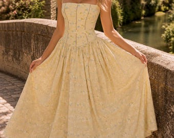 May include: A pale yellow midi dress featuring a delicate blue and white floral pattern, a fitted bodice with thin spaghetti straps, a square neckline, and a full, pleated skirt that falls below the knees, paired with simple tan slide sandals.