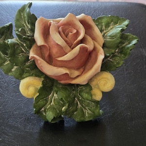 May include: A decorative wall plaque featuring a central dusty rose-colored flower with layered petals, surrounded by textured dark green leaves and two small, pale yellow scroll-like accents at the base, crafted with a dimensional relief design.