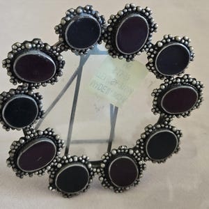 May include: Circular silver-toned metal photo frame with a ring of alternating black and burgundy oval-shaped stones. The frame has a decorative beaded edge and a clear glass or plastic insert.