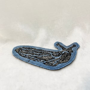 May include: A blue and black fabric patch in the shape of a whale. The whale is outlined in blue and has a black and silver glitter design.