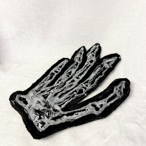 May include: A black fabric patch with a white skeleton hand design. The hand is outstretched with fingers pointing upwards.