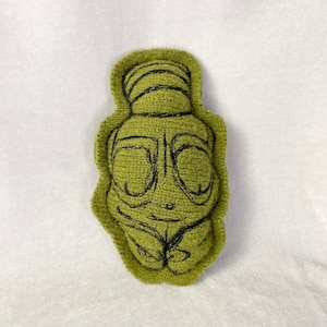 May include: A green plush toy shaped like a person with a black embroidered face and hair. The toy has a simple design and is made of soft, fuzzy fabric.