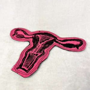 May include: A pink fabric patch with a black outline in the shape of a uterus.