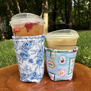 Pode incluir: Duas mangas de tecido para copos envolvendo copos de plástico para bebidas geladas. A manga mais alta apresenta um padrão floral azul e branco. A mais curta tem um fundo azul claro com um padrão repetido de ilustrações de vida marinha.