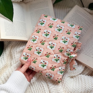 May include: A pink quilted book sleeve with a repeating pattern of strawberries and white cloud-like shapes. The book cover has a matching tie closure. The background includes open books and a white knitted blanket.