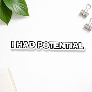 May include: A white sticker with the text "I HAD POTENTIAL" in a bold, black font. The sticker has a black outline. The sticker is on a white surface, with binder clips and a notebook visible.