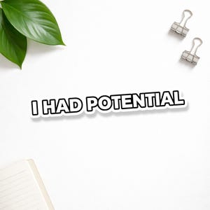 May include: A white sticker with the text "I HAD POTENTIAL" in bold, black letters with a white outline. The sticker is on a white surface, with green leaves, binder clips, and a notepad in the background.