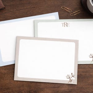 May include: Three stationery notepads with colored borders in blue, green, and beige, arranged on a wooden surface. One notepad has the initials "TRE" printed on it. A silver pen, paper clips, and a cup of coffee are also visible.