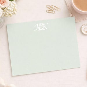 Può includere: Un foglio di carta da lettere verde chiaro con le iniziali "NRK" in scrittura bianca. La scena comprende una penna rosa, un taccuino rosa, una tazza di caffè e un piattino con un anello. Sono presenti anche un mazzo di fiori e un nastro.