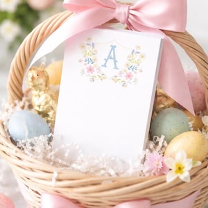 May include: A woven Easter basket containing pastel eggs, paper, and a notepad. The notepad displays a floral wreath design with the letter "A". A pink satin ribbon is tied around the basket, which is filled with Easter decorations.