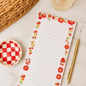 Könnte beinhalten: Ein liniertes Notizbuch mit Tomatenrand, ein goldener Stift und eine rot-weiß karierte Schale liegen auf einer Marmoroberfläche. Ein Glas Wasser mit einer Zitronenscheibe und ein Weidenkorb sind ebenfalls im Bild.