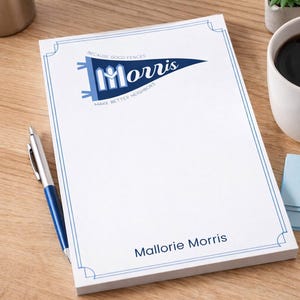 May include: White notepad with a blue border and the name "Morris" in a pennant design. The notepad also includes the text "Because good fences make better neighbors" and the name "Mallorie Morris". A blue and silver pen rests on the wooden surface.