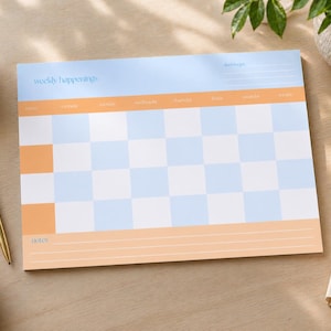 May include: A weekly planner pad with a light blue and orange color scheme. The pad features a checkerboard pattern and the words "weekly happenings." A gold pen and a cup of coffee are next to the planner.