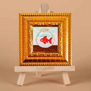 May include: A small, framed painting of a red goldfish in a glass bowl. The painting is set against a light blue and pink background, with a brown base. The frame is ornate gold, and the painting is displayed on a small wooden easel. The text "TulipArtsIN" is visible.