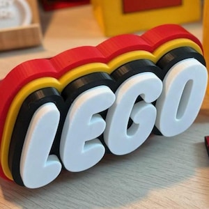 Può includere: Un'insegna tridimensionale che presenta la parola LEGO in grassetto con lettere maiuscole bianche. Le lettere sono montate su una base a strati composta da una sezione centrale nera, un bordo giallo e un bordo esterno rosso.