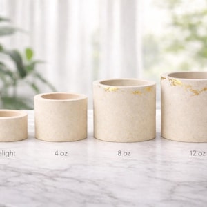 Puede incluir: Cuatro portavelas cilíndricos beige de diferentes tamaños, etiquetados como "Tealight", "4 oz", "8 oz" y "12 oz". Los dos portavelas más grandes tienen detalles de pan de oro. Los portavelas están sobre una superficie de mármol.
