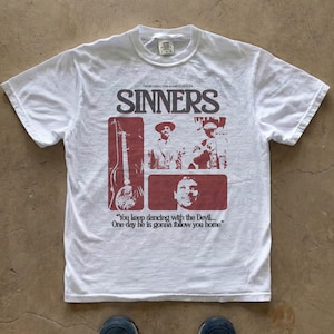 Puede incluir: Camiseta blanca con un diseño gráfico con la palabra "SINNERS" e imágenes de músicos y una guitarra en un rectángulo rojo. El texto "You keep dancing with the Devil... One day he is gonna follow you home" también está impreso.