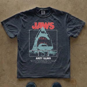 Könnte beinhalten: Dunkelgraues T-Shirt mit dem Wort "JAWS" in Rot über einer Hai-Grafik. Unter dem Hai steht der Text "AMITY ISLAND" und weiterer Text. Das T-Shirt ist aus weichem Material. Das T-Shirt hat eine klassische Passform.