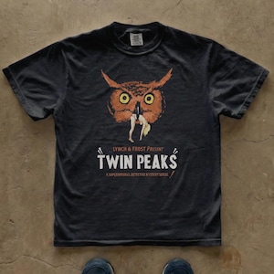 Może przedstawiać: Ciemnoszara koszulka Twin Peaks z ilustracją sowy trzymającej kobietę. Tekst "LYNCH & FROST PRESENT TWIN PEAKS" i "A SUPERNATURAL DETECTIVE MYSTERY SERIAL" są poniżej. Koszulka leży płasko.