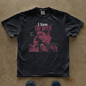 May include: A charcoal grey short-sleeve t-shirt featuring a graphic print from the film I Saw the Devil. The design includes a portrait of a man, three smaller inset images, and the text: I Saw the Devil and You can't kill me. You'll become just like me.