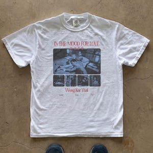 May include: A white short-sleeve t-shirt featuring a graphic print from the film In the Mood for Love by Wong Kar Wai. The design includes a large black and white scene of two people, smaller film stills, and red text reading In the Mood for Love.