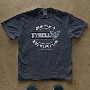Może przedstawiać: Ciemnoszary t-shirt z krótkim rękawem z białym nadrukiem graficznym logo Tyrell Corporation, symbolem sowy i japońskim tekstem. Projekt zawiera słowa Genetic Replicants oraz More Human Than Human.