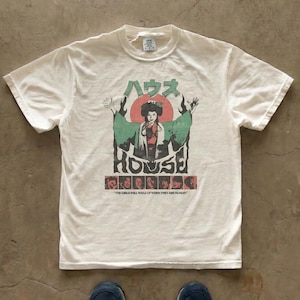 Könnte beinhalten: Ein cremefarbenes Kurzarm-T-Shirt mit einem Grafikdruck einer Person in einem grünen Gewand, darüber der japanische Text "ハウス" und darunter das englische Wort "HOUSE". Eine Reihe kleiner Porträts und der Text "THE GIRLS WILL WAKE UP WHEN THEY ARE HUNGRY" sind unten abgebildet.