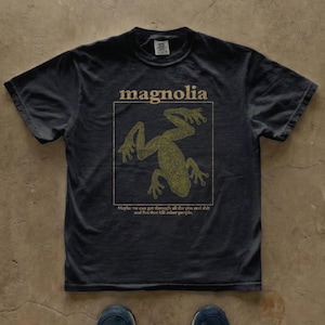 May include: A charcoal gray short-sleeve t-shirt featuring a gold-toned graphic of a frog inside a square frame. Above the frame is the word "magnolia" and below it is the text "Maybe we can get through all the piss and shit and lies that kill other people."