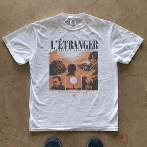 May include: A white short-sleeve t-shirt featuring a graphic print related to the novel L'Étranger by Albert Camus. The design includes the title, author, and illustrations of scenes with the text C'est à cause du soleil and a small sun icon.