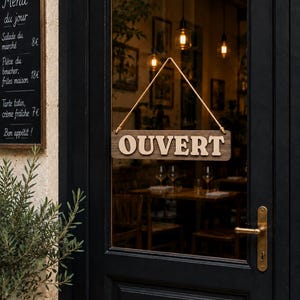 Peut inclure: Une enseigne rectangulaire en bois suspendue par une corde sur une porte noire. L'enseigne porte le mot OUVERT en lettres capitales claires et en relief sur un fond en bois sombre. La porte possède une poignée en laiton et une vitre.