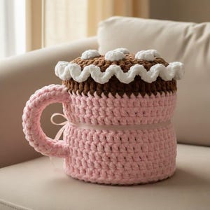 May include: A pink crocheted mug cosy with a handle and a cake-shaped lid. The lid is brown with white accents, resembling frosting. A pink ribbon is tied around the mug. The cosy is designed to fit over a mug, adding a decorative touch.