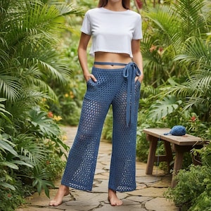 Op de afbeelding: Blauwe gehaakte broek met een wikkelband in de taille en zakken. De broek is gemaakt van een losse, open geweven stof. Een witte crop top is ook zichtbaar. De broek wordt buiten in een tuin getoond.