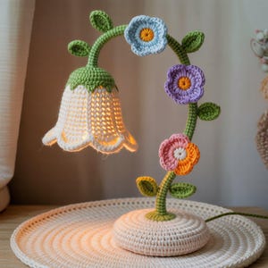 以下が含まれることがあります： 手作りの花の形をしたかぎ針編みのランプ。白いベル型のシェード、緑色の茎、青、紫、ピンクのかぎ針編みのカラフルな花が特徴です。ランプは丸いクリーム色のベースに置かれています。