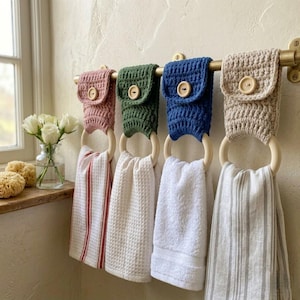 May include: A brass towel rack holds four crocheted towel holders in pink, green, blue, and beige. Each holder has a wooden ring and a button closure. The towels hanging from the rings are striped, waffle-textured, and solid white.